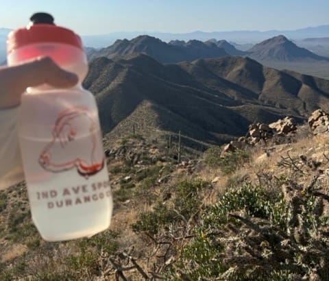 a hiker in an extremely hot desert holds a water bottle out in a cactus covered scene