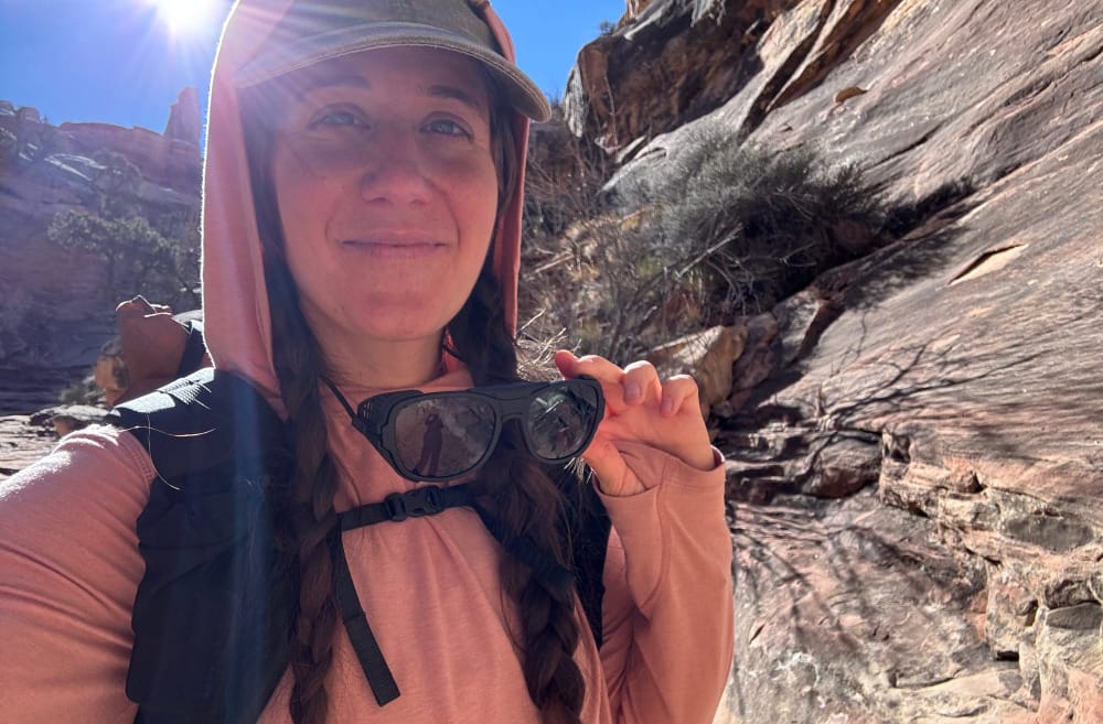 A woman shows her ombraz teton sunglasses on a bright day hiking in a deep canyon in the southwest