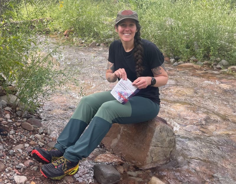 a hiker wears a pair of the best hiking pants for women of 2025 while having a snack along a creek