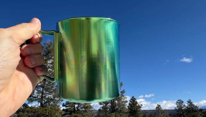 a hiker holds the Ti-Double 450 Anodized Mug By Snow Peak in a forested blue sky scene